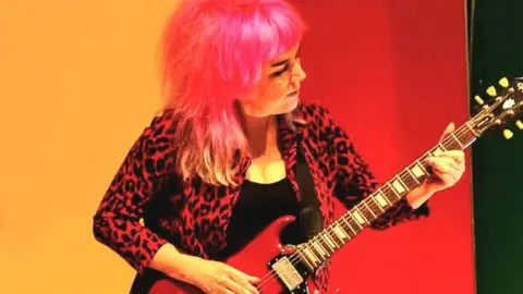 Dan Lord A woman with pink hair plays guitar. She is wearing a red leopard print jacket and a black T shirt.