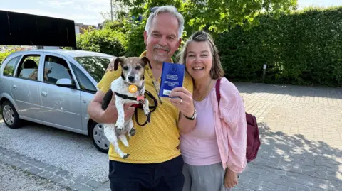 David and Suzanne Nicholl with their small white and brown Jack Russell terrier dog, Lily. Mr Nicholl has white hair and a goatee and is wearing a mustard colour T-shirt and black trousers. He is holding the dog under his right arm and the dog's blue Belgian passport in his left hand towards the camera . Mrs Nicholl's has bobbed brown hair with the front section up in a small quiff. She is wearing a pink T-shirt, pink cardigan and grey trousers. She has a maroon bag on her left shoulder and her right arm around Mr Nicholl's. 