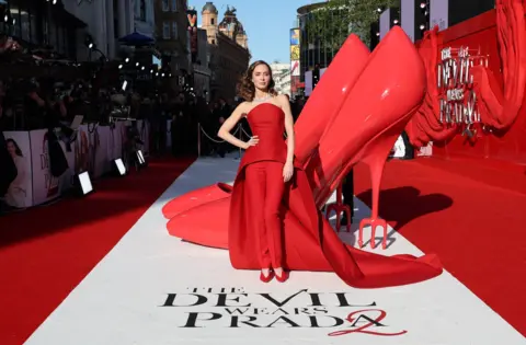 Getty Images Emily Blunt poses at the red carpet premiere. She is standing next to a sculpture of a giant pair of red stilettos. 