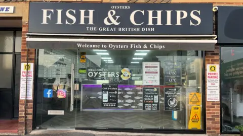 Adrian Harms / BBC A fish and chip shop with a black sign above it and a glass exterior. The shop sign reads "Oysters / Fish & Chips / The Great British Dish". The glass window of the shops has a number of small signs reading "welcome to oysters" as well as prices and social media logos.