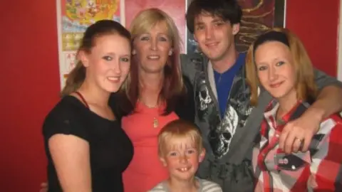 Rosevear Family A photo of a family embracing each other. Two women in their 20s stand either side of a young man and a slightly older woman, while a small boy smiles in front of them.