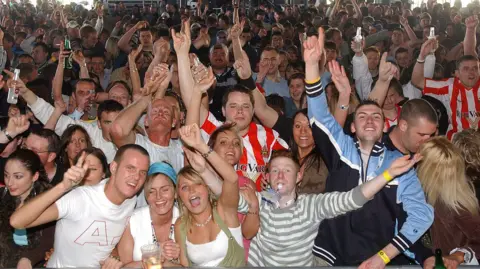 A picture taken from the stage of a crowd of people singing and dancing with arms in the air. Several are wearing red and white-striped Sunderland shirts, in one corner there is a couple kissing. A man at the front with short ginger hair is blowing a whistle, there are two young women with blonde hair cheering next to him and many people are clutching plastic drink glasses and bottles.