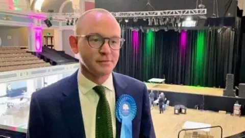 A picture of Andrew Snowden taken on a balcony above an election counting hall, there are green and purple lights against a black curtain hanging above the stage in the background, there are seats to his left. He is wearing a blue suit, with a green tie and a Conservative rosette.