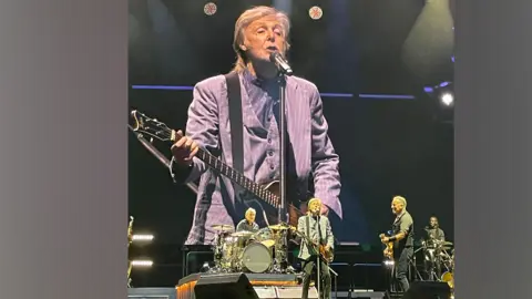 bruce2023tour /X Sir Paul McCartney wearing a grey blazer, stands in front of a microphone, holding a guitar, on a huge stage. Bruce Springsteen is to his left, smiling at him, as he holds a guitar. A man is on the drums behind them.