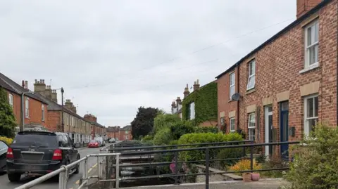 North Kesteven District Council A row of terraced houses on a narrow street. There is a waterway in front with access to the houses via a walkway. There are cars parked on the road.