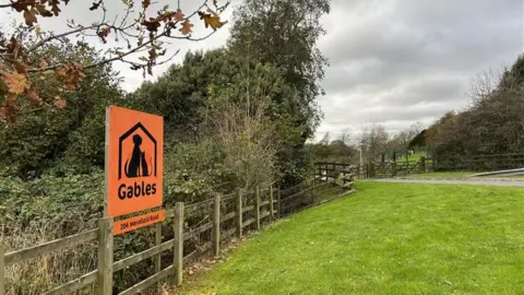 An orange Gables sign on a grassy area. The sign is attached to a wooden fence and there are bushes and trees behind the fence.
