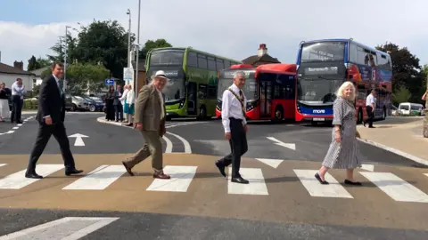 Four dignitaries - three men in suits, including the major with a chain and woman in a striped dress - walk across the new zebra crossing at Salisbury Railway Station, in the style of a famous Beatles album cover