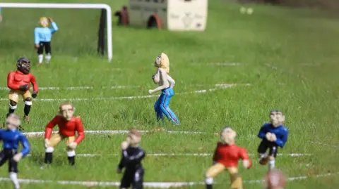 A close up of a football pitch with miniature model players. A figure of a topless blonde-haired woman can be seen running across the pitch.