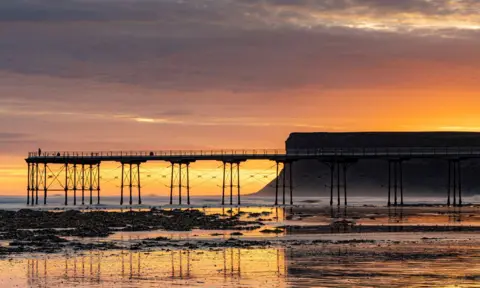 Andrew Sturdy A pier juts out into the sea with a cliff and orange sunrise beyond, all reflected in the wet sand in the foreground.