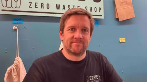 Graham Rutherford in a black t-shirt standing in front of a zero waste shop sign