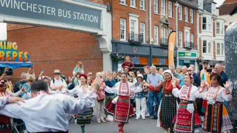 Adrienne Photography A group of women in traditional costumes dance in the street outside Winchester Bus Station with a large crowd watching on.