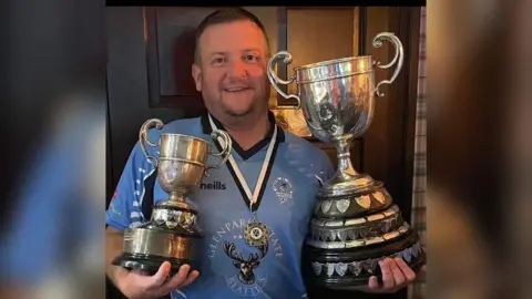 Sion Mills Cricket Club A cricketer, in a blue uniform, holds two trophies, one in each arm. A medal is around his neck and he is smiling.