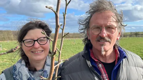 Former dairy farmers Debra and Tom Willoughby standing next to an apple tree sapling