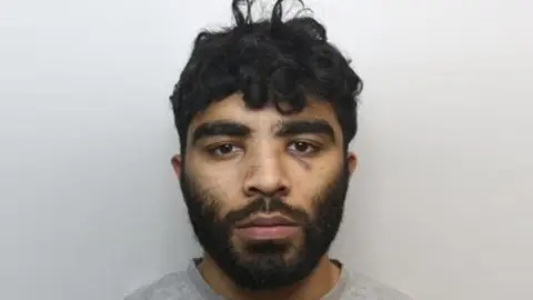 A police mugshot of Hassan Jhangur. He has dark facial hair and dark, slightly curly hair.