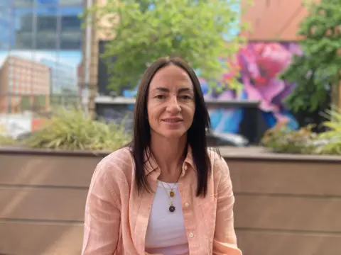 A woman with dark hair looks at the camera. She is wearing a pale pink shirt and white vest. 