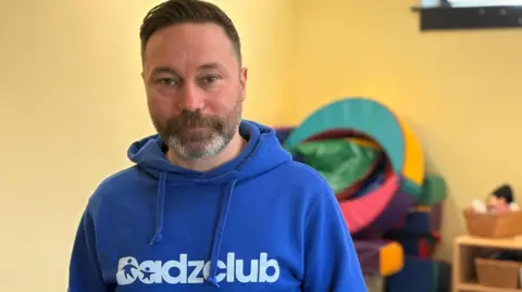 Matt Foster wearing a blue jumper with the logo Dadzclub. He is standing in a room full of toys, one of the venues for his community groups.