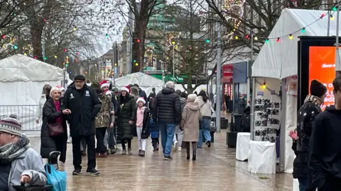 BBC Crowds walk down a soggy street in Hull. Coloured lights hang from the trees. Stalls run down the side of the street.