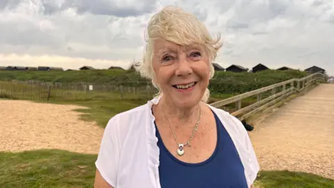 Contributed Jan Etherington smiles at the camera. She is wearing a blue top, white blouse and silver necklace and has silver hair. She is standing by a bridge with beach huts visible behind her.