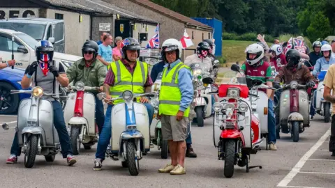 Andrew David A group of around 30 people are all on vespa style scooters, many of them have union jack flags on them. They look like they are about to set off on a ride. 