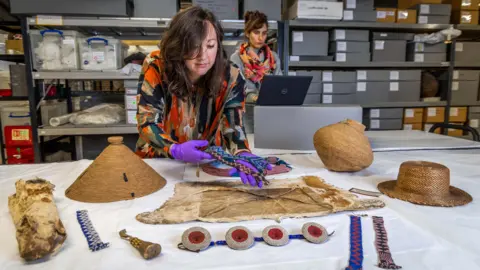 The image shows a woman wearing purple gloves looking at and holding artefacts on a table; behind her is a woman typing on a laptop.