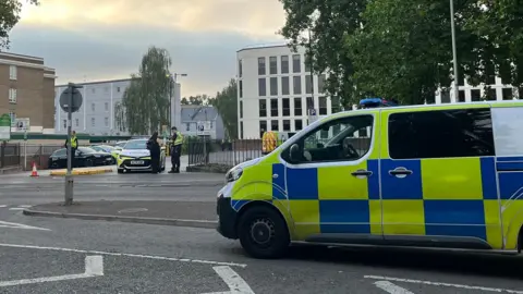 A police van is parked on a road opposite the entrance to Rodney Road car park, where a police van is parked. Two officers can be seen at the entrance, with one speaking to someone dressed in black. There are trees and buildings surrounding the car park, which has a few civilian cars parked in it.
