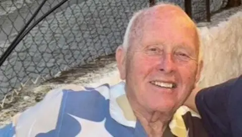 A man with short grey hair leans back, smiling at the camera. He is wearing a patterned blue, yellow and cream shirt