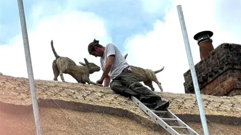 Chris Fellows Chris Fellows working on a thatched rood. His legs are resting on a metal ladder he had used to get to the top. There are two straw hounds on the roof beside him. It's a cloudy day.