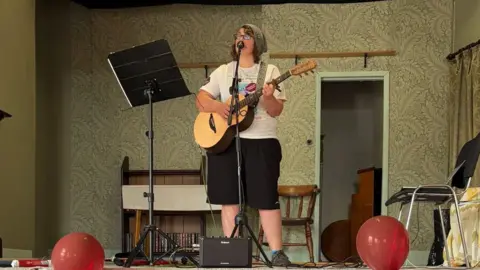 Supplied A woman wearing a white T-shirt and black shorts performing on stage. She has an acoustic guitar and is singing into a microphone. 