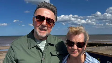 Crispin Rolfe/BBC Robert and Lynne Tomlinson pay their respects. Robert is wearing sunglasses and a green shirt. Lynne is also wearing sunglasses and a striped blue and white top.