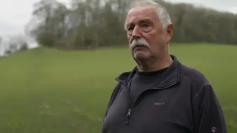 A man with short white hair and a white moustache is pictured in a green field with trees behind him. 