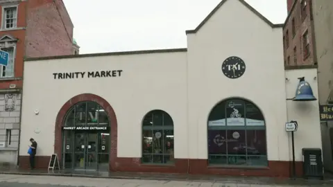 An early 20th century market hall building with an arched entrance and two matching windows. It is painted cream with red brick details. A round black clock face doubles as a sign with the letters "T M" printed on it. Another sign reads "Trinity Market". To the right, a large blue bell advertises the Blue Bell pub.
