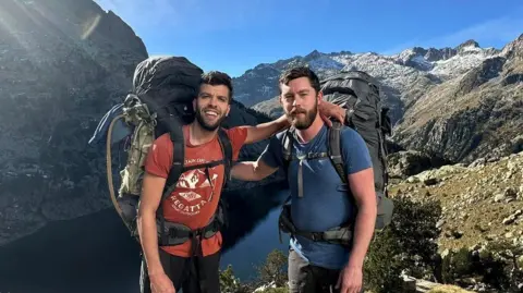 File photo of Aziz Ziriat (left) and Sam Harris in shorts and T-shirts wearing large hiking backpacks and stood on top of a hilltop overlooking a gorge with their arms on each others' shoulders.