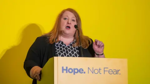 Pacemaker Naomi Long is standing behind a podium which says Hope not fear. She is speaking into a microphone and is wearing a black and white top, with a black long sleeved cardigan over it. Behind her is a yellow wall. 