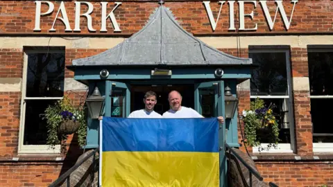 Two men are holding a Ukrainian flag and standing outside a hotel.