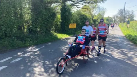 Karen Rees Gareth Rees sits in his adapted race wheelchair being pushed along a rural road by his team of three runners behind him. They are wearing red and white race bibs. A yellow road closure sign is on the side of the road behind him