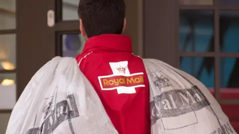 A Royal Mail worker in a bright red jacket carries a large sack over both shoulders