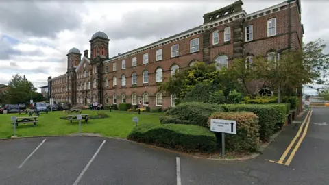 Google A Google Street View screenshot of Skiddaw House, part of the Fusehill campus. The building has a main central body with a pitched roof, a tower at either side and a clock on the higher part of the facade. There are two large wings over three floors on both sides of the central body. There are picnic tables on a patch of grass outside the building and a parking area with cars.