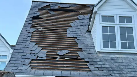The Lihou Charity LBG A close up of a rood where tiles have fallen off. There wood on the roof is exposed. To the right is a window which appears undamaged. The sky is blue.
