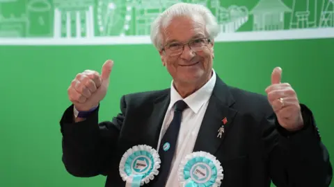 PA Media A man with white hair and glasses is wearing a smart black suit and tie. He is putting both thumbs up and is wearing two turquoise rosettes.