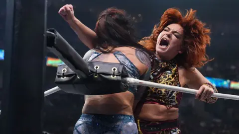 AEW Jamie Hayter delivers a clothesline wrestling move to her opponent, who is pressed against the turnbuckles in the ring