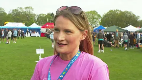 A woman standing outdoors on a grassy field. She is facing slightly to one side rather than directly at the camera. Sunglasses are resting on top of her head, and a blue lanyard with visible printed text and logos hangs around her neck over a pink short-sleeved T‑shirt.
