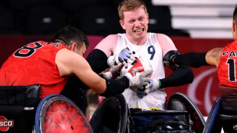 Getty Images A man with ginger hair and a white vest top in a black wheelchair holding a white ball while a man with dark hair and a red vest top tries to take it off him