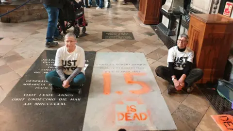 Two Just Stop Oil are seen sitting on the floor in Westminster Abbey at each side of Darwin's marble nave wearing white t-shirts with the group's name on it. "1.5 is dead" is painted on in large orange font.