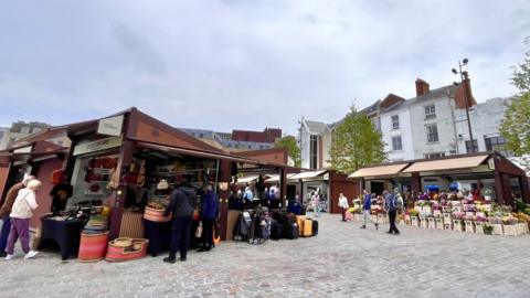 An outdoor market scene features several wooden stalls arranged on a cobblestone square. One stall on the left displays various baskets and colorful woven goods, while farther back stalls offer flowers and other items to shoppers. People are casually walking and browsing the market under an overcast sky, with white and light-colored buildings surrounding the square in the background.
