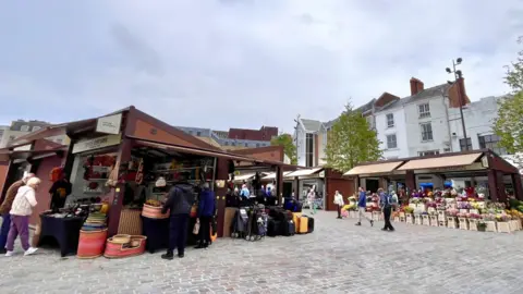 Martin Giles/BBC  Brown-coloured market stalls selling woven baskets, luggage and flowers while people walk around on cobbled paving 