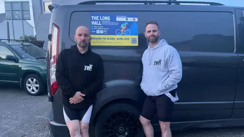 JODIE CANNELL Adrian a bald man in a black jumper and Darren a man with brown hair and stubble wearing a grey jumper stand next to a black van with a poster that says the long hall.