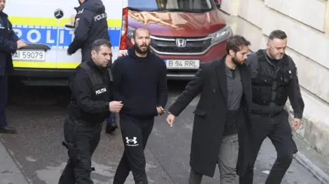 Getty Images Police accompanying Andrew and Tristan Tate, who are handcuffed to one another, to court in Bucharest