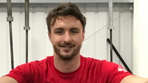 Aaron Gulliver in a gym, wearing a red T-shirt and smiling at the camera.