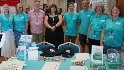 Eight people are smiling and standing behind a table with goodies and leaflets to give away, including black baseball caps with 200 on them. Most of the team are wearing pale blue T-shirts with 200 on them.