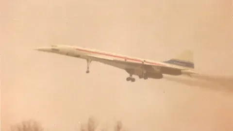 The Concorde prototype is seen taking off into the air. It is white with a red and a blue stripe.